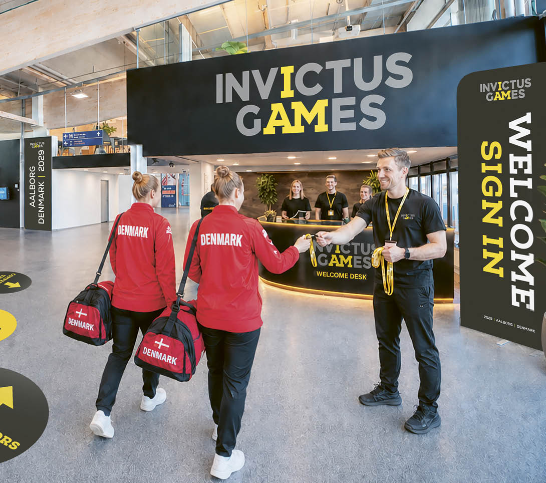 A group of people are walking in a building with a sign that says Invictus Games. Description generated by AI